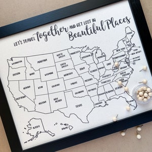 May include: A white canvas map of the United States with black outlines of each state. The text "Let's Travel Together and Get Lost in Beautiful Places" is at the top of the map. There are small white push pins placed on the map.