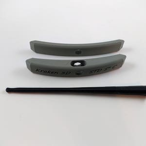 Puede incluir: Dos piezas de plástico gris con las palabras "Kraken 3D" y "STD 5" impresas en ellas. También se incluye una varilla de plástico negro.