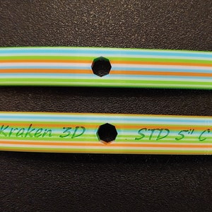 Puede incluir: Dos espaciadores de plástico impresos en 3D de colores vivos con un agujero hexagonal en el centro. El espaciador superior tiene rayas verdes, naranjas, blancas y azules. El espaciador inferior tiene el texto "Kraken 3D . STD 5" C" impreso en él.