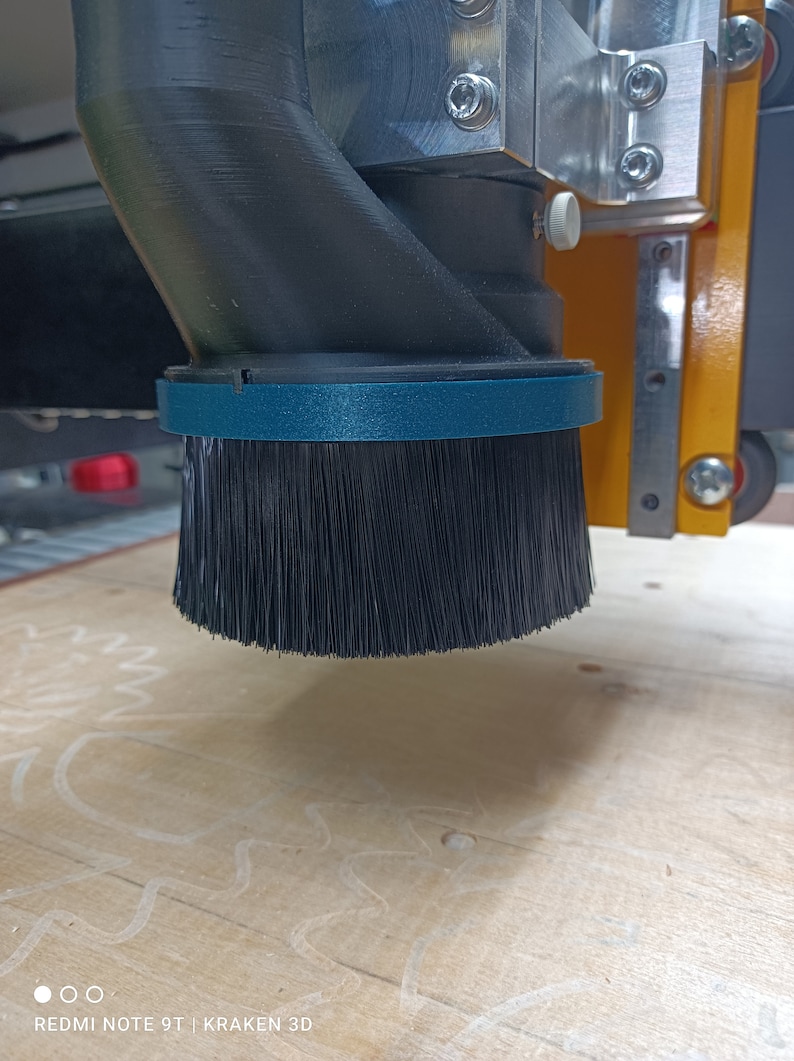 May include: A close-up of a black dust collection brush attached to a CNC machine. The brush is made of soft bristles and is designed to collect dust and debris from the work surface.