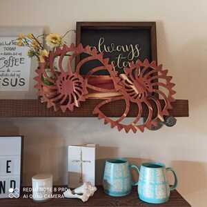 Könnte beinhalten: Zwei hölzerne Wanddekorationen in Form von Sonnenblumen mit einem braunen Holzregal im Hintergrund. Das Regal hat auch ein Schild mit der Aufschrift "always stay" und ein weißes Schild mit dem Text "ALL NE TODAY a little bit of coffee & whole lot of ESUS".