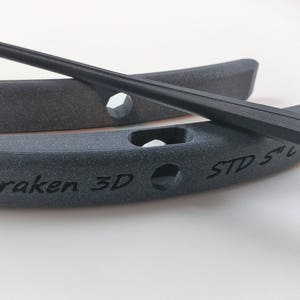 Puede incluir: Dos soportes negros impresos en 3D para una espada impresa en 3D. Los soportes son curvos y tienen el texto "Kraken 3D" y "STD 5" impreso en ellos.