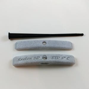 Puede incluir: Dos presas de escalada de plástico gris, curvas, con el texto "Kraken 3D" y "STD 5" C" impreso en ellas. También se muestra una herramienta de goma negra.