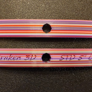 Puede incluir: Dos bridas de cable de plástico de colores con un agujero en el centro. La brida superior no tiene texto. La brida inferior tiene el texto "Kraken 3D STD 5" impreso en ella.