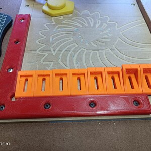 May include: A close-up of a red and orange plastic woodworking jig with multiple slots. The jig is made up of two pieces, one red and one orange. The orange piece has multiple slots that are used to hold the wood in place while it is being cut.