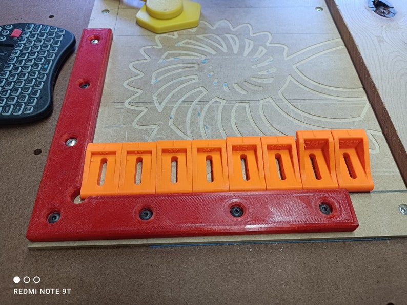 May include: A close-up of a red and orange plastic woodworking jig with multiple slots. The jig is made up of two pieces, one red and one orange. The orange piece has multiple slots that are used to hold the wood in place while it is being cut.
