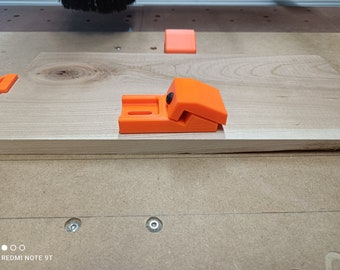Puede incluir: Abrazaderas de plástico naranja para trabajar la madera, utilizadas para sujetar piezas de madera juntas. Las abrazaderas están sobre una superficie de madera.