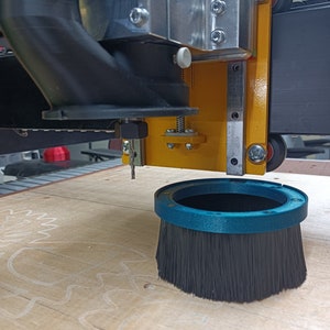 May include: A close-up of a CNC router with a black dust collection hose and a blue brush attachment. The router is positioned over a piece of wood with a white pencil drawing of a gear.
