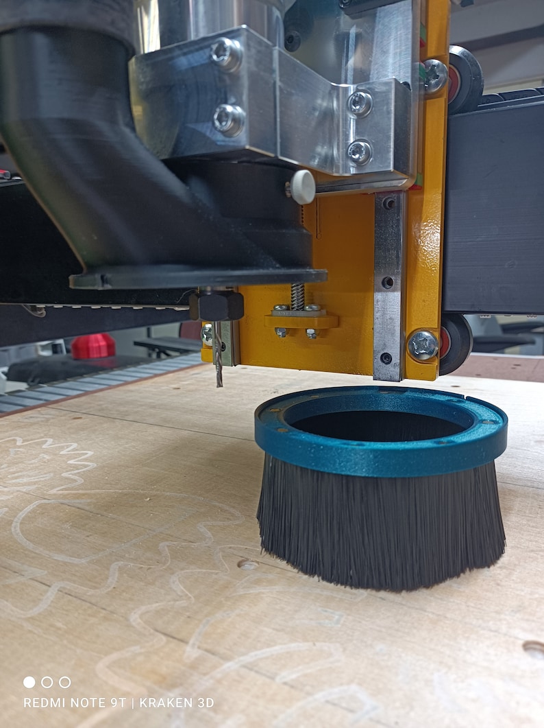 May include: A close-up of a CNC router with a black dust collection hose and a blue brush attachment. The router is positioned over a piece of wood with a white pencil drawing of a gear.