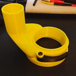 May include: A yellow plastic 3D printed router jig with a black clamp. The jig has a round opening and a long, curved spout.