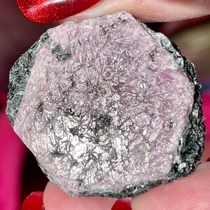 K&ouml;nnte beinhalten: Ein roher rosafarbener Rubin-Edelstein mit schwarzen Einschl&uuml;ssen. Der Rubin ist ein roher, ungeschliffener Stein mit sechseckiger Form.