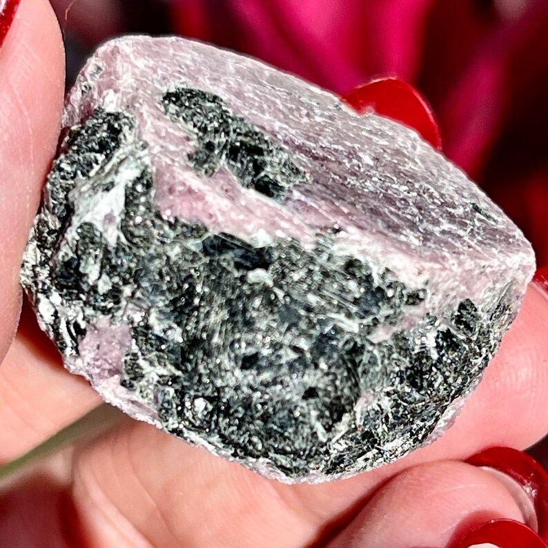 K&ouml;nnte beinhalten: Ein rauer, rosa-schwarzer Edelstein mit einer nat&uuml;rlichen, unregelm&auml;&szlig;igen Form. Der Edelstein hat eine raue, strukturierte Oberfl&auml;che und eine tiefrosa Farbe.