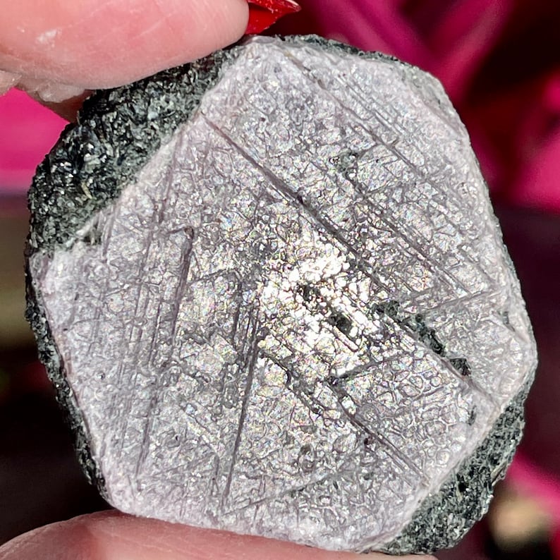 K&ouml;nnte beinhalten: Ein roher, ungeschliffener, graulilafarbener Edelstein mit sechseckiger Form und einer rauen, strukturierten Oberfl&auml;che. Der Edelstein ist von einer dunkelgrauen Matrix umgeben.