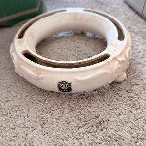May include: Off-white ceramic decorative ring with a floral design on the sides and a small emblem. The ring has a circular shape with a hollow center and is resting on a light-colored carpet.