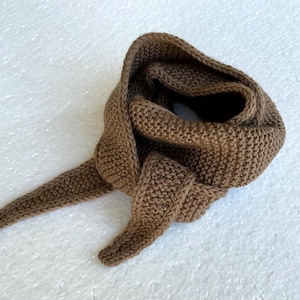 Könnte beinhalten: Ein brauner Strickschal mit strukturierter Optik. Der Schal ist gefaltet und drapiert, mit einem langen, spitz zulaufenden Ende, das sich nach links erstreckt. Der Strickstoff hat eine warme, erdige braune Farbe, passend für kaltes Wetter.