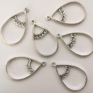 Pode incluir: Seis pingentes de joias em formato de gota feitos de metal prateado. Cada pingente apresenta uma moldura externa texturizada e uma barra horizontal interna decorativa com um design de corda trançada e pequenos laços pendentes. Cada peça tem um anel.