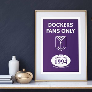 Puede incluir: Una impresión enmarcada con un fondo morado y texto blanco que dice "DOCKERS FANS ONLY". La impresión también presenta un logotipo de ancla blanco y el texto "ESTABLISHED 1994 FREEMANTLE FOOTBALL CLUB".