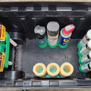 Puede incluir: Una caja de herramientas negra llena de suministros de pintura. Incluye pinceles, botes de pintura en aerosol, tubos de masilla y recambios de rodillos de pintura. Los pinceles están en un soporte verde y los recambios de rodillos son amarillos.