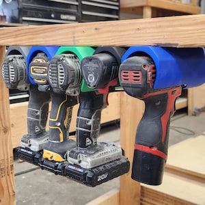 May include: A set of five plastic power tool mounts in blue, green, and black colours, attached to the underside of a wooden workbench. Each mount holds a cordless drill or driver, with various battery packs attached to the base of each tool.