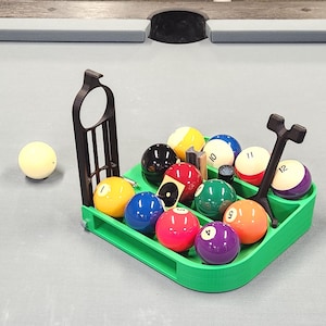 Puede incluir: Un juego completo de bolas de billar, numeradas del 1 al 15, en un soporte verde, con una bola blanca y un puente negro. Las bolas están dispuestas sobre una mesa de billar gris.