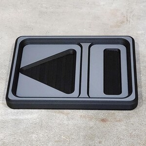 May include: A rectangular, black tray with a gray border and gray play and pause symbols. The play symbol is a triangle pointing left, and the pause symbol is a vertical rectangle.