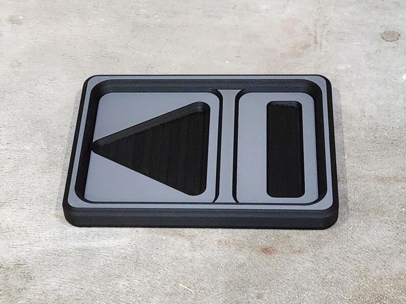 May include: A rectangular, black tray with a gray border and gray play and pause symbols. The play symbol is a triangle pointing left, and the pause symbol is a vertical rectangle.
