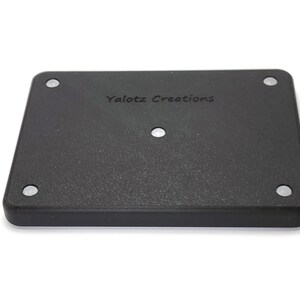 May include: A black rectangular object with rounded corners and the text "Yalotz Creations" printed on the surface. The object has four small, silver-colored circular accents in each corner and one in the center.