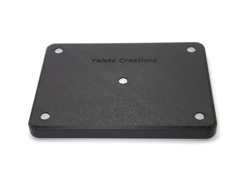 May include: A black rectangular object with rounded corners and the text "Yalotz Creations" printed on the surface. The object has four small, silver-colored circular accents in each corner and one in the center.