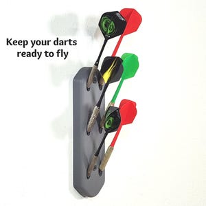 May include: A grey wall-mounted dart holder with three slots holding three darts with red and green flights. The text "Keep your darts ready to fly" is printed to the left of the holder. The darts feature metallic barrels and black shafts.