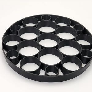 May include: Black circular object with multiple cylindrical holes of varying sizes. The object is made of a matte black material and sits on a white surface. The design features a series of interconnected, circular openings.