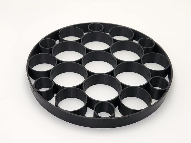 May include: Black circular object with multiple cylindrical holes of varying sizes. The object is made of a matte black material and sits on a white surface. The design features a series of interconnected, circular openings.