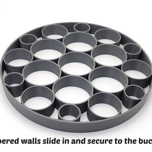 May include: A gray, circular object with multiple cylindrical openings. The text "Tapered walls slide in and secure to the bucket" is visible at the bottom of the image. The object appears to be made of plastic or a similar material.
