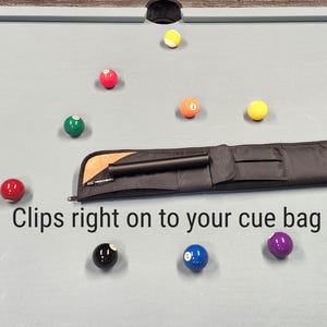 May include: A black cue case accessory with a cylindrical shape and a metal clip, positioned on a light gray pool table surface surrounded by scattered colorful billiard balls. The text "Clips right on to your cue bag" is printed below the item.