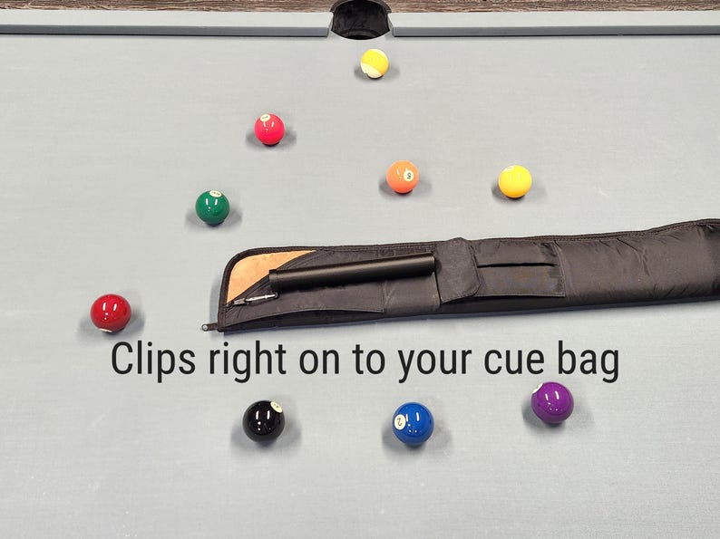 May include: A black cue case accessory with a cylindrical shape and a metal clip, positioned on a light gray pool table surface surrounded by scattered colorful billiard balls. The text "Clips right on to your cue bag" is printed below the item.