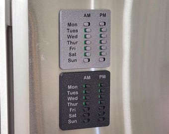 May include: Two rectangular magnetic pill organizers, one silver and one black, featuring vertical columns labeled AM and PM with rows for each day of the week from Mon to Sun. Each day has a sliding switch with a green indicator for tracking medication.