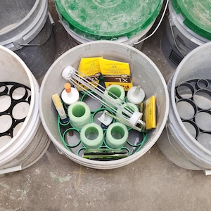 May include: An overhead shot of several gray buckets. One bucket contains various green and yellow construction supplies, including rollers and sponges. Other buckets contain black and white circular inserts.