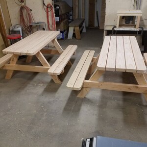 May include: Two unfinished wooden picnic tables with benches. The tables are made of natural wood and have a simple design.