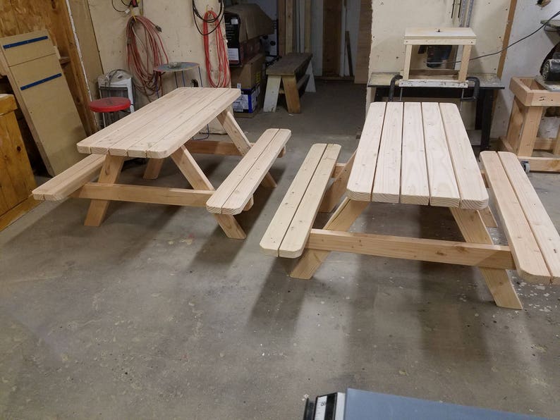 May include: Two unfinished wooden picnic tables with benches. The tables are made of natural wood and have a simple design.