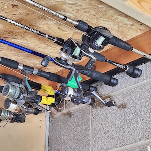 May include: A black wall-mounted fishing rod rack holding four fishing rods with attached reels. The rods feature various colors including silver, blue, and black, with visible brand text such as Vengeance and Silvermax on the rod blanks.