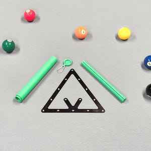 May include: A black triangular billiard ball rack, two green cylindrical tubes, and a small green circular cap with a metal carabiner clip, surrounded by several colorful numbered billiard balls on a gray surface.