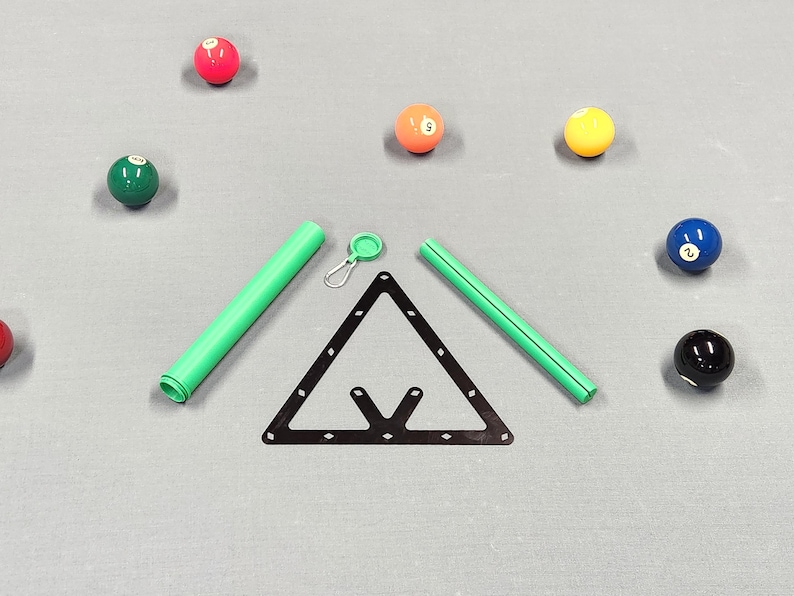 May include: A black triangular billiard ball rack, two green cylindrical tubes, and a small green circular cap with a metal carabiner clip, surrounded by several colorful numbered billiard balls on a gray surface.