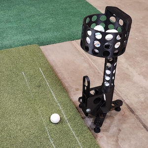 Op de afbeelding: Een zwarte golfbaldispenser met een ronde mand vol golfballen. De dispenser staat op een statief en staat op een groene grasmat. Een enkele golfbal ligt op het gras.