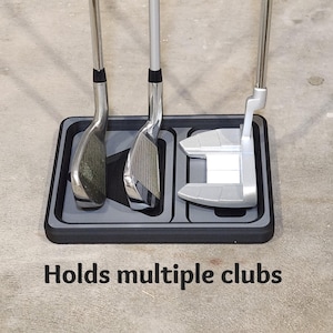 May include: A black and gray golf club holder with three golf clubs. The holder has a rectangular shape with three slots to hold the clubs. The text "Holds multiple clubs" is printed on the surface.