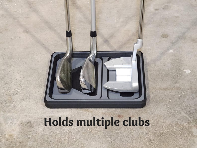 May include: A black and gray golf club holder with three golf clubs. The holder has a rectangular shape with three slots to hold the clubs. The text "Holds multiple clubs" is printed on the surface.
