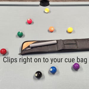 May include: A black cue bag with a grey cylindrical accessory attached to the exterior, featuring a metal clip. The text "Clips right on to your cue bag" is printed below the bag, which is surrounded by various colored billiard balls.
