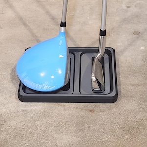 May include: A golf club driver with a light blue head and a silver shaft rests in a black rectangular tray. A silver golf club iron is also in the tray. The tray is on a concrete surface.