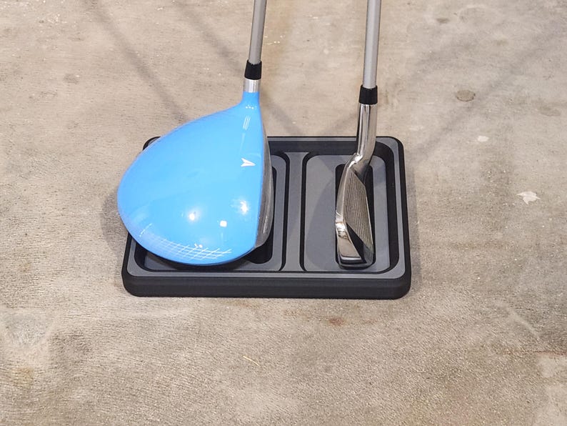 May include: A golf club driver with a light blue head and a silver shaft rests in a black rectangular tray. A silver golf club iron is also in the tray. The tray is on a concrete surface.