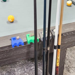 May include: A set of four plastic pool cue holders in blue, green, gray, and black, each featuring a slot for coins. These holders are attached to the edge of a pool table, supporting four vertical pool cues with varying shaft colors and designs.