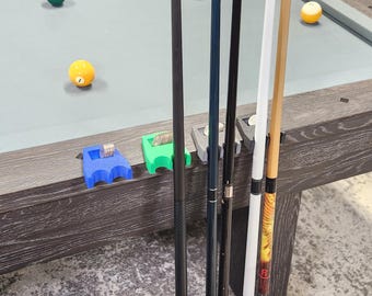 Könnte beinhalten: Ein Set aus vier Billard-Queue-Haltern aus Kunststoff in Blau, Grün, Grau und Schwarz, jeweils mit einem Schlitz für Münzen. Die Halter sind am Rand eines Billardtisches befestigt und stützen vier vertikale Billard-Queues mit verschiedenen Designs.