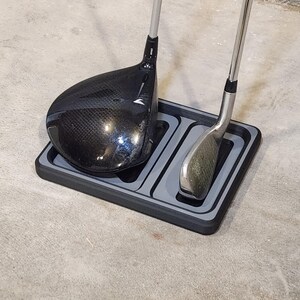 May include: A black and gray golf club organizer holding a black driver and a silver iron. The organizer has two compartments and is sitting on a concrete surface.